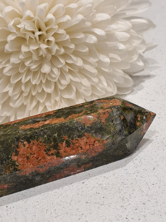 Natural unakite crystal tower in red and green, photographed with a chrysanthemum flower in the background. 