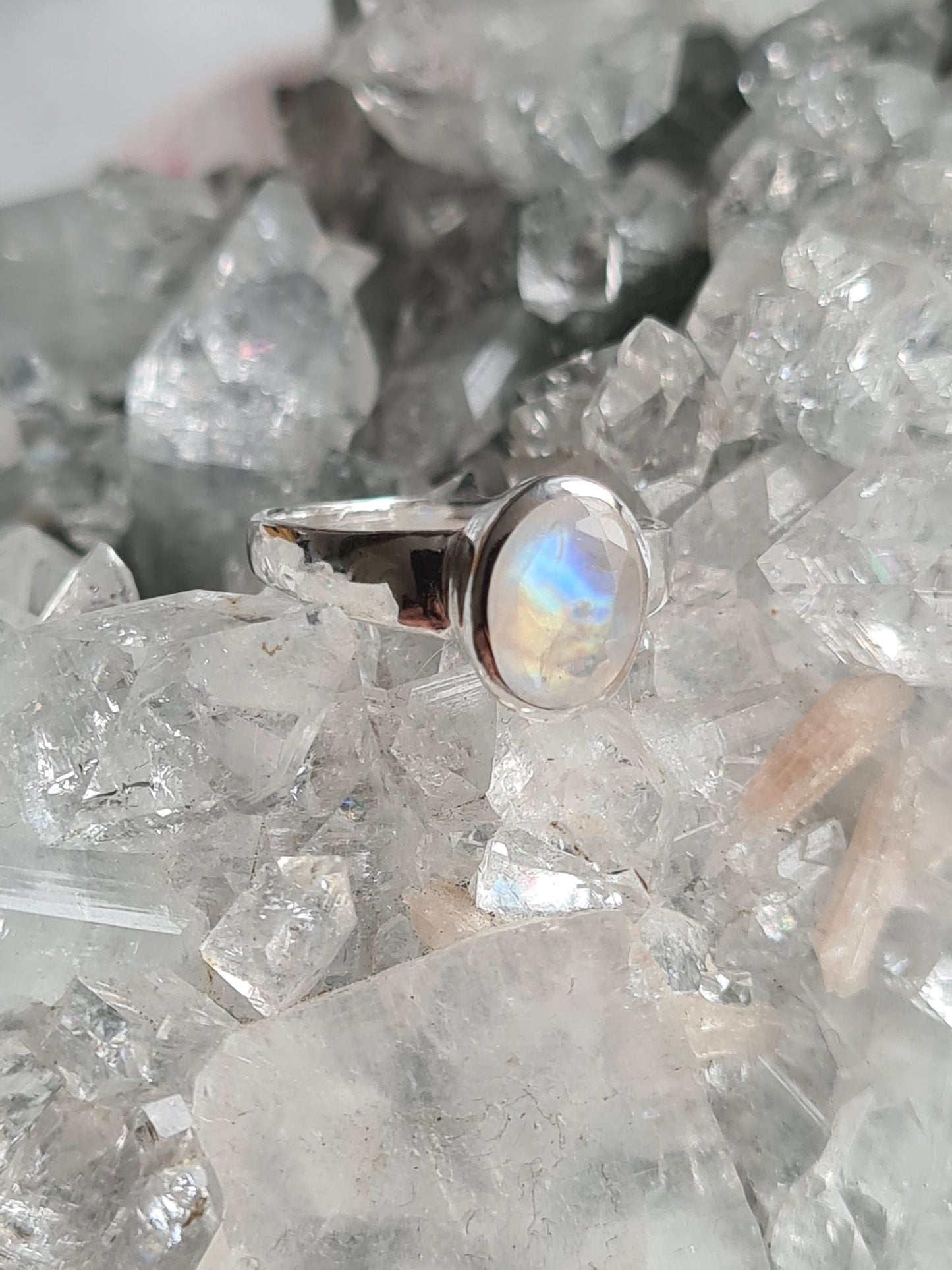 An oval faceted rainbow moonstone set ring in sterling silver. Shown on crystal cluster background. 