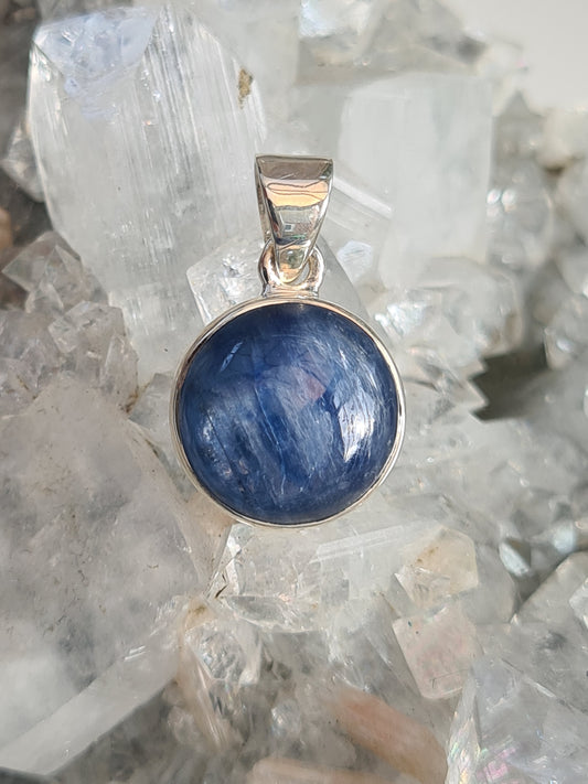 A round blue kyanite single stone pendant in sterling silver. Rubover set bezel. 
Photographed on a white apophyllite cluster crystal.