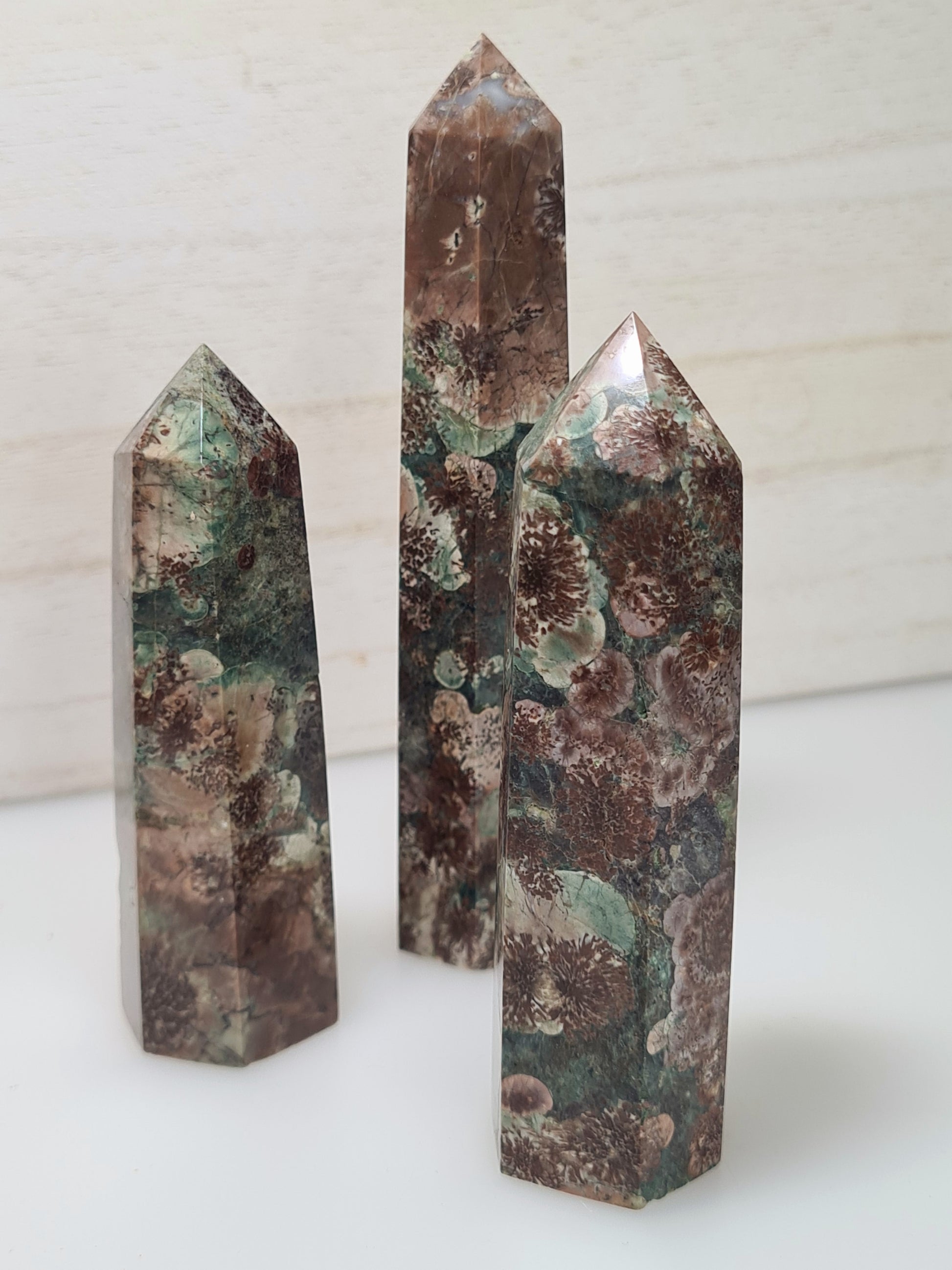 Three Natural Plum Blossom Jasper Towers, body colour green with reddish brown Rhyolite flower looking inclusions. Photographed on a white wooden background.