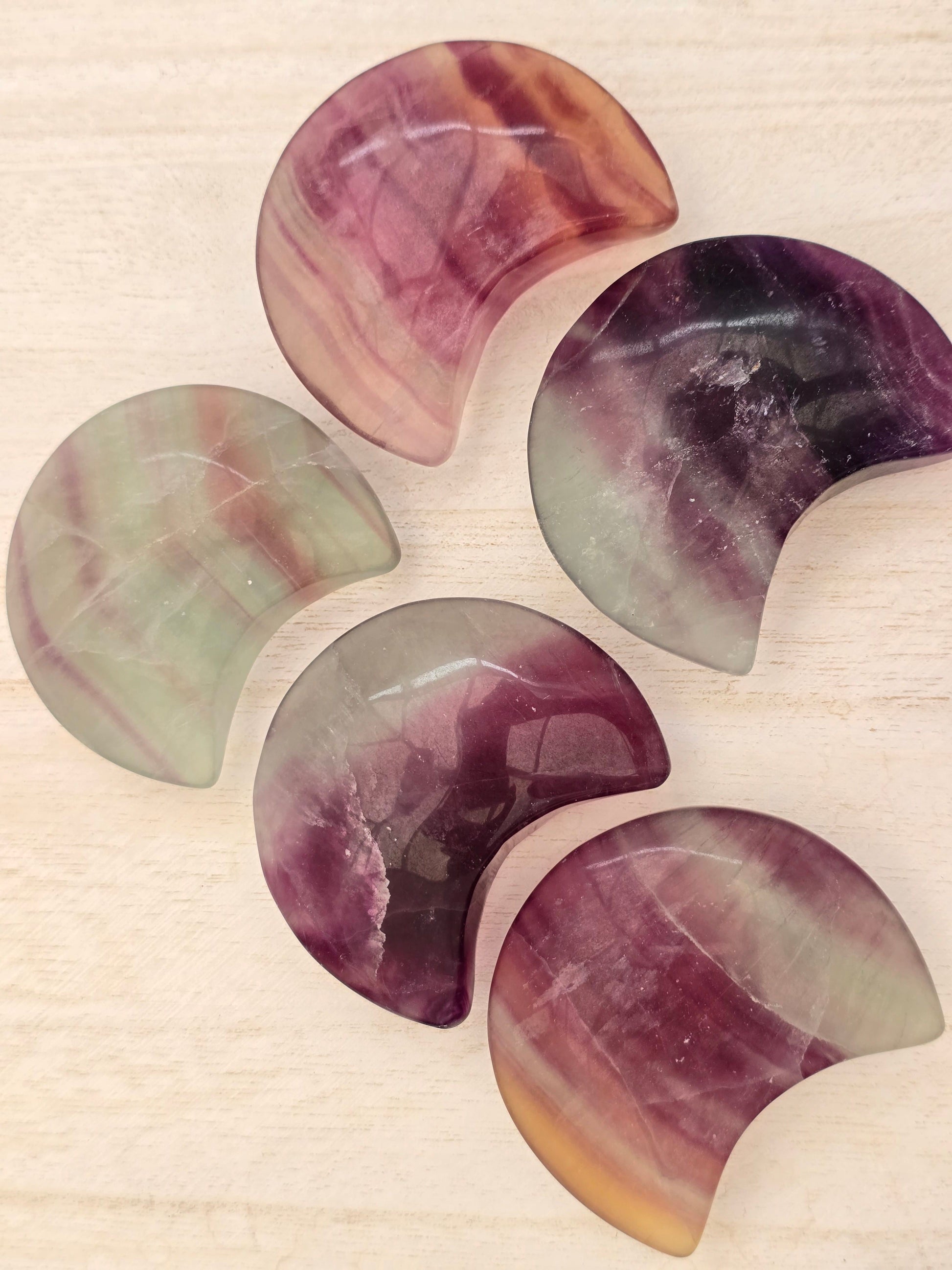 Five natural rainbow fluorite large crescent moon carvings. Green and purple are the dominant colours. 
Photographed on a wooden background.