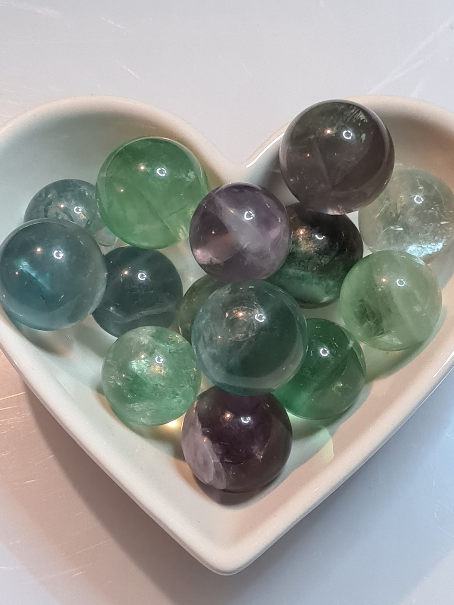 White heart shaoed bowl filled with mulitcoloured rainbow fluorite small spheres, colours of green and purple found most frequently