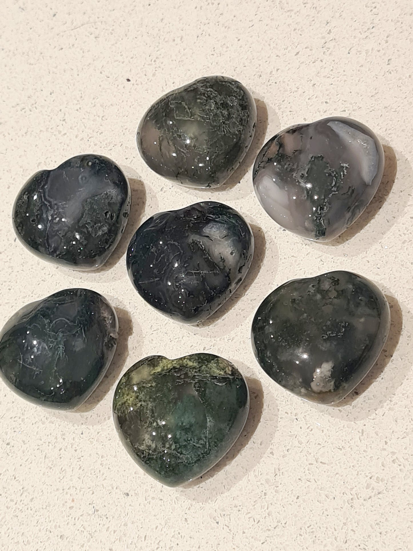 Seven Smooth Moss Agate Small Heart Crystal Carvings set out in a daisy formation on a white background