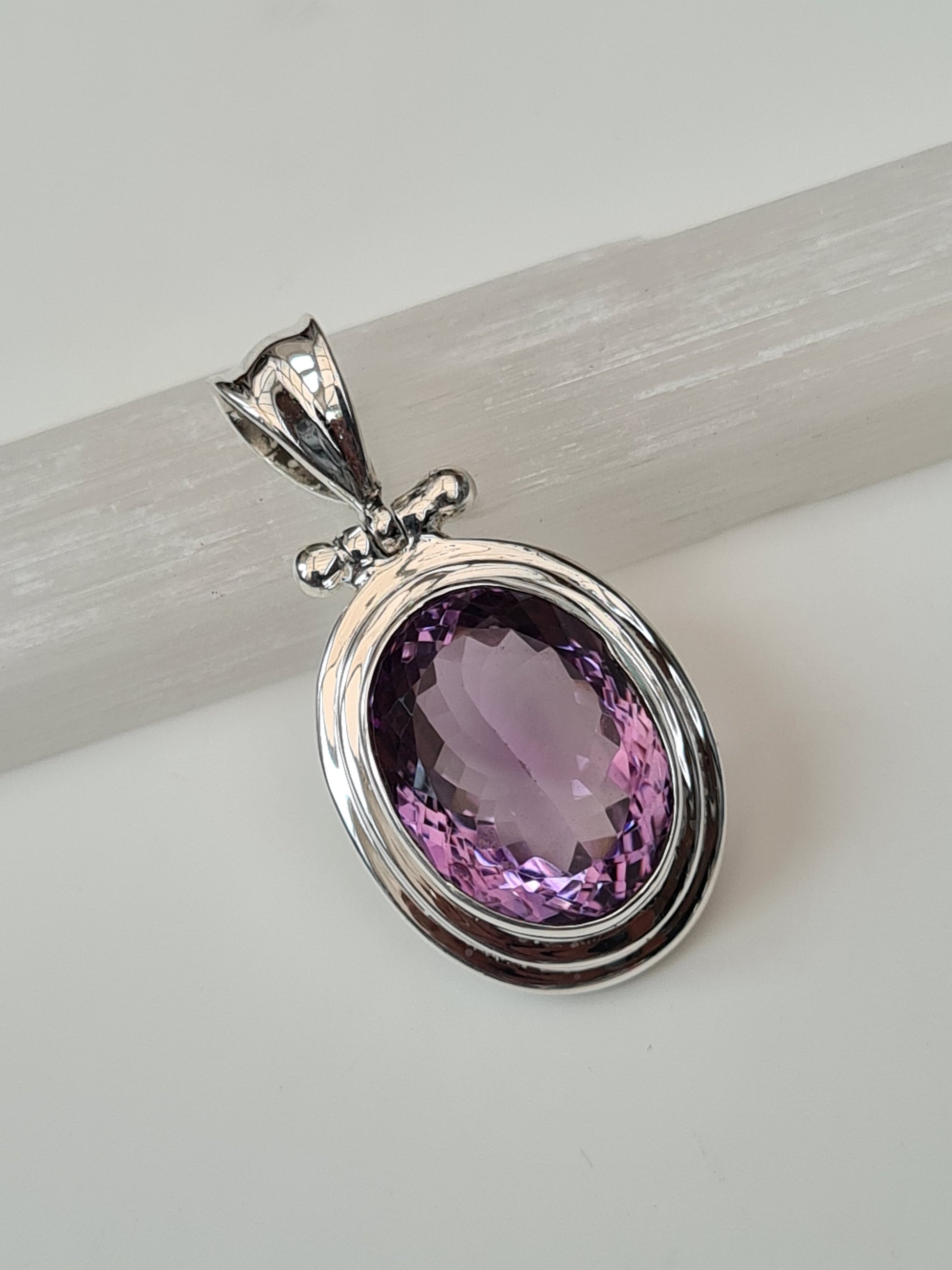 An oval shaped faceted amethyst, rubover set to a polished 2 row frame with hinged grooved bail. 925 sterling silver