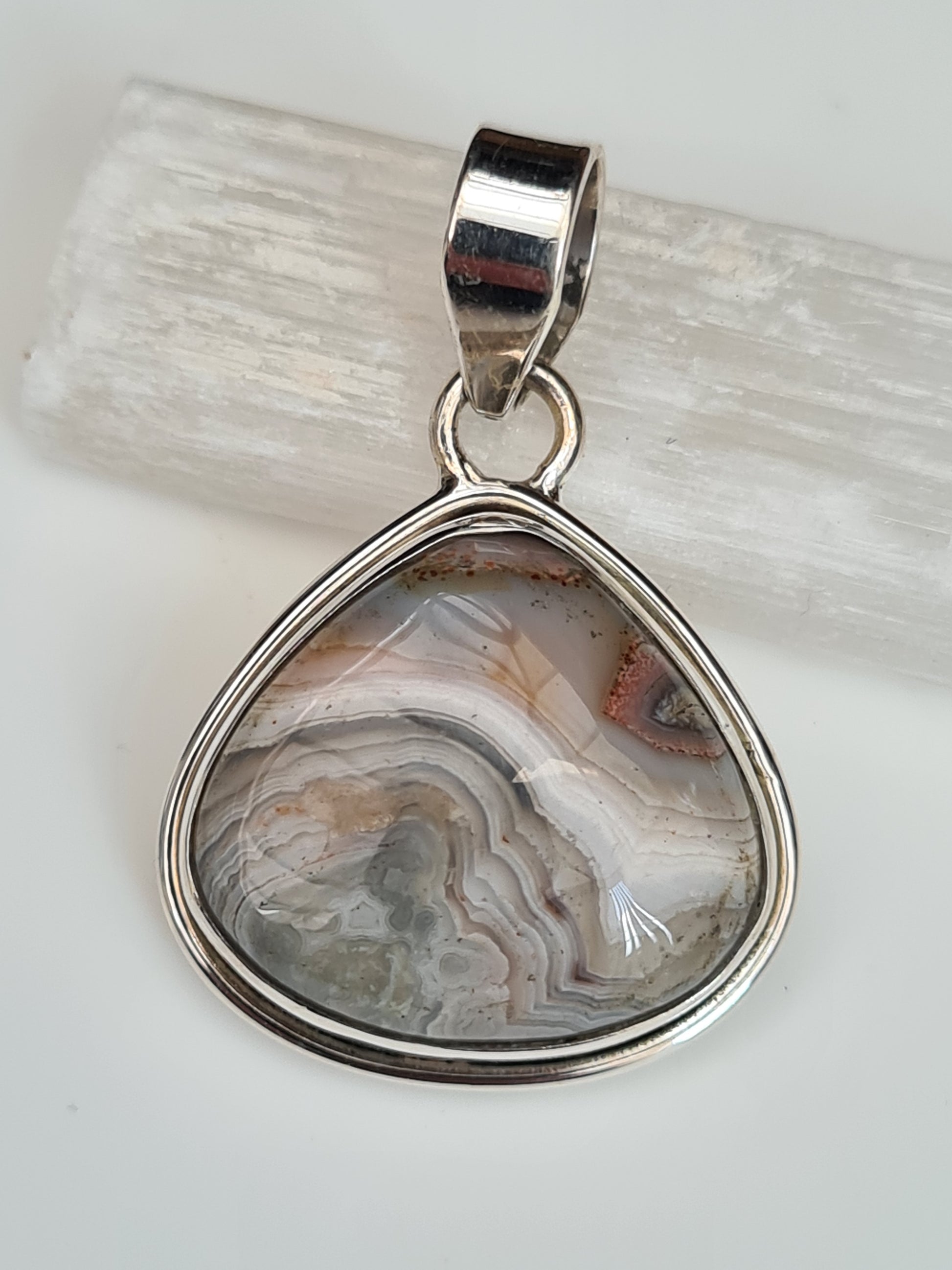 A Pear Shaped Crazy Lace Agate Cabochon Set Pendant in Sterling Silver. Colours of grey, white and red.
Photographed on a white selenite stick and background.