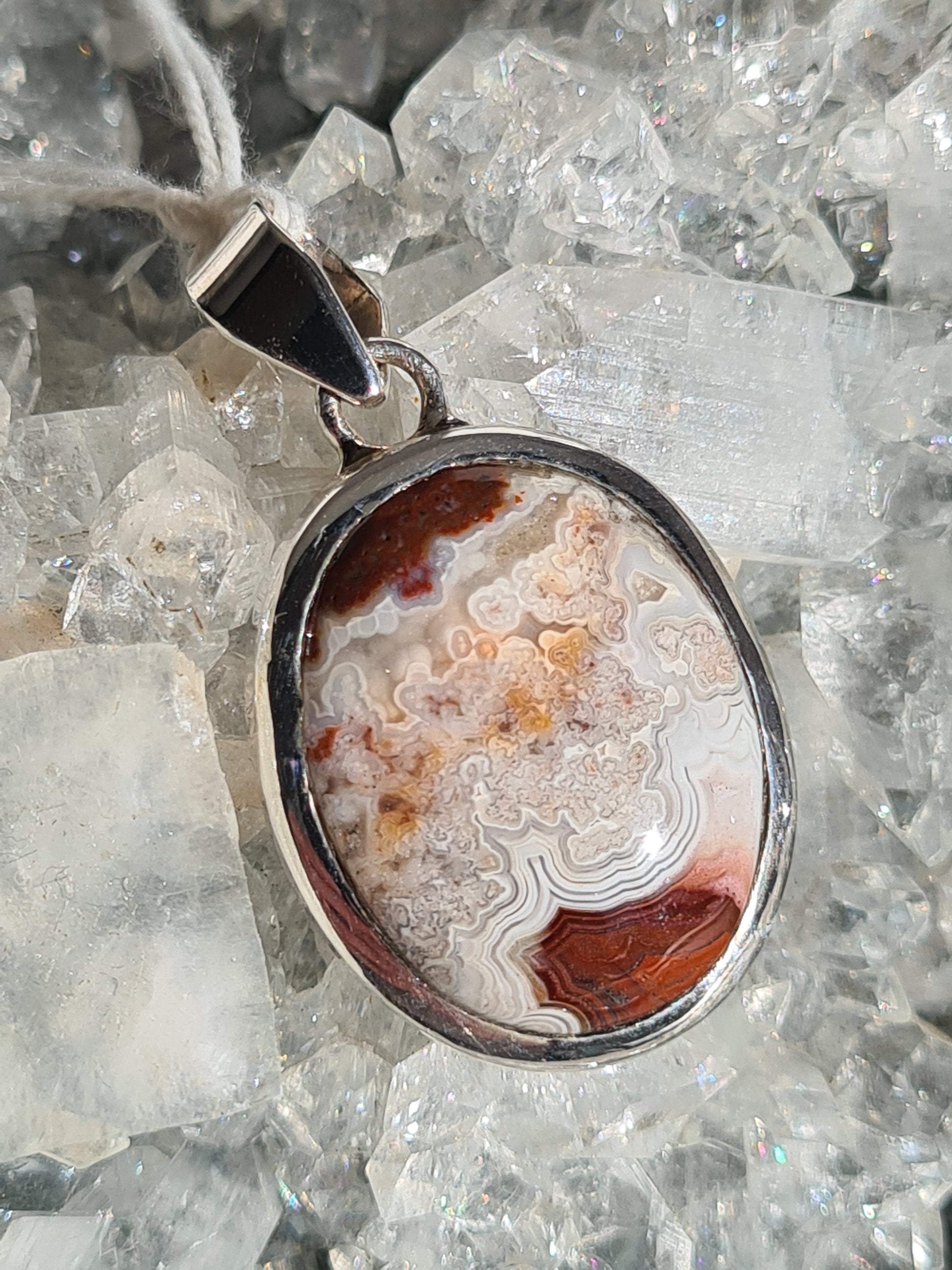 A Mexican crazy lace agate set pendant in sterling silver. Large oval, with red, white and grey banding.
Photographed on an apophyllite cluster background. 