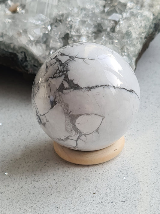 Natural White Howlite Sphere, approx. 50mm diameter. White bodycolour with grey magnesite veins. Photographed on a wooden Sphere stand with a raw cluster in the background 