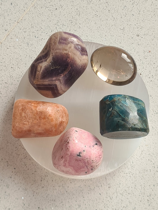 A selenite charging plate with five intuitively chosen tumbles. This set contains apatite, sunstone, rhodonite, smoky quartz and dream amethyst.
